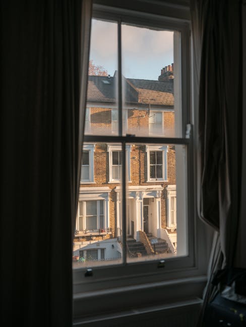 A view through a double-glazed window with white wooden framing, showing the exterior of a row of Victorian-style terraced houses with brick facades and white window frames. The window has dark curtains pulled back on either side, revealing a partly cloudy sky with soft daylight. The inner window sill appears clean and free of dust or clutter, and the window reflects a tidy indoor environment. This scene highlights the importance of maintenance and cleanliness in domestic settings, consistent with the surface cleaning and deep cleaning services offered by Maida Vale Cleaners during end of tenancy cleans. The overall image emphasizes clarity and brightness, enhancing the perception of a well-maintained home or property.
