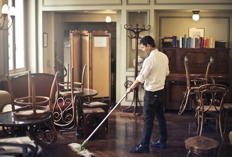 A professional cleaner from Maida Vale Cleaners wearing a white shirt and dark trousers is performing surface cleaning in a living room, using a mop on a polished wooden floor. The room features a mix of wooden and upholstered furniture, including a round table with chairs stacked upside down on top and a wooden cabinet with books and decorative items. Large windows allow natural light to illuminate the space, highlighting the cleanliness and shine of the wooden surfaces. The room appears tidy, with no visible dust or debris, and the surfaces are well-maintained. The setting suggests a thorough domestic cleaning process aimed at maintaining hygienic, spotless surfaces, emphasizing the company's focus on high-quality deep cleaning and sanitisation services.