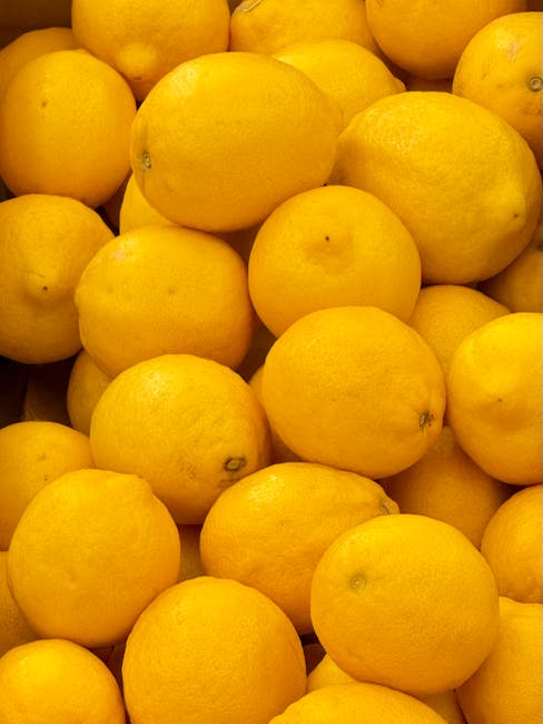 A close-up view of a large pile of fresh, bright yellow lemons with smooth surfaces and some small imperfections, arranged on a surface in a well-lit setting. The vivid yellow color contrasts with the subtle shadows, highlighting the natural texture of the citrus fruit. This image emphasizes the freshness and clean appearance of the lemons, which could be part of a domestic or commercial setting related to food preparation or display, in line with surface cleaning and hygiene standards promoted by Maida Vale Cleaners. The lemons are in focus with a sharp detail, capturing the round shape and slight variations in size, with no visible dirt or blemishes, indicating freshness and cleanliness.