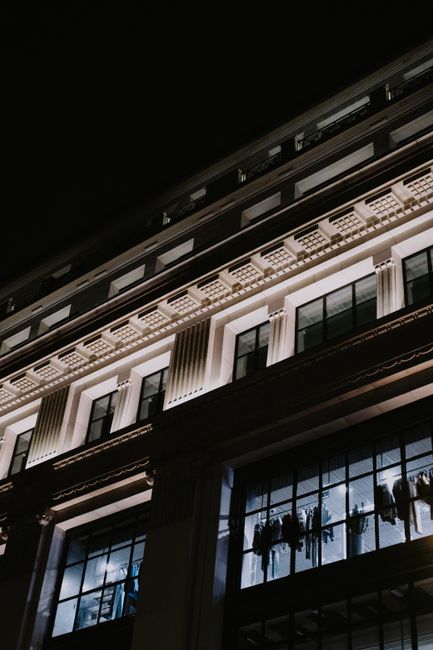 A nighttime exterior view of a multi-story building with classical architectural details, including decorative cornices and large window panes. The windows reflect interior lighting and display hanging plants and dining furniture inside. The building's façade is illuminated to highlight its textured surfaces, and the dark sky contrasts with the lit architecture. This image showcases the clean, well-maintained appearance of an urban commercial property, as part of the services offered by Maida Vale Cleaners for surface cleaning and regular maintenance of shop fronts and office exteriors.