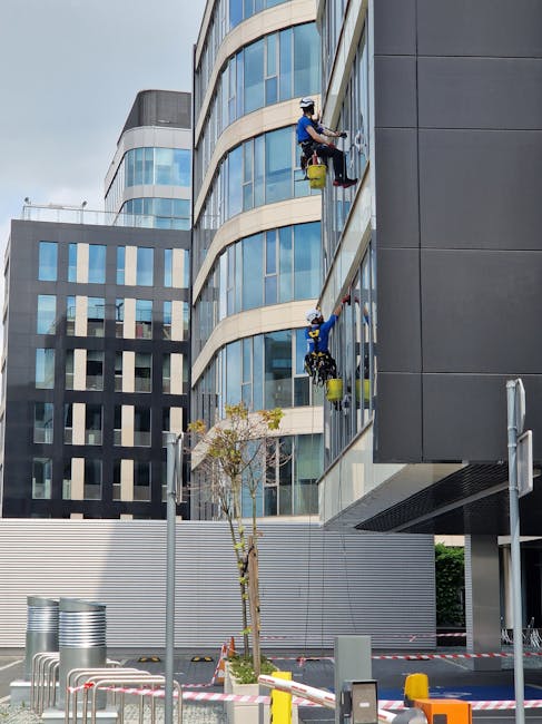 Two window cleaners wearing safety helmets, harnesses, and blue uniforms are working on the exterior glass facade of a modern multi-story office building in Maida Vale. They are seated on suspended platforms and using squeegees and cloths to clean the large, curved glass windows. The building features curved architectural lines with reflective blue glass panels and dark grey cladding sections. The street level shows a small tree, street furniture, and some barriers, with an overcast sky overhead. This scene illustrates professional commercial cleaning services provided by Maida Vale Cleaners, focusing on high-rise window cleaning and facade maintenance.