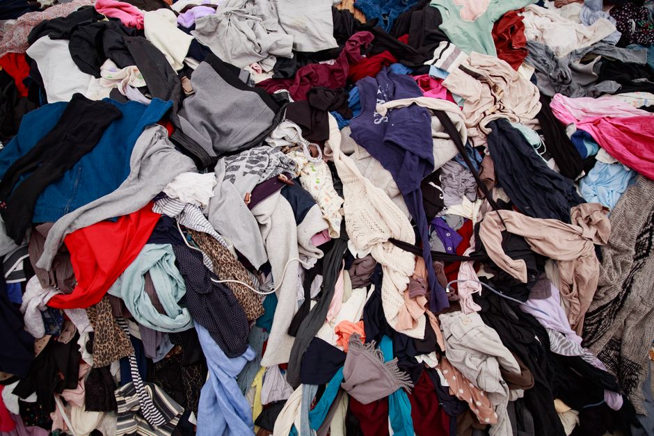 A large pile of mixed laundry consisting of various clothing items including t-shirts, pants, dresses, and accessories, all scattered and folded in a disorderly manner on a flat surface. The clothing