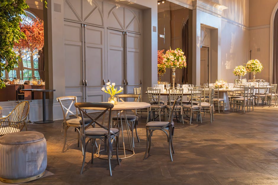An elegant event space with polished wooden floors and high ceilings, featuring round tables decorated with tall glass vases filled with fresh pink and white flowers, accompanied by silver Chiavari chairs with cushioned seats. The room is well-lit with natural light from large windows, and the surfaces, including the tabletops and floors, appear spotless and well-maintained. A small side table holds a yellow floral arrangement, and soft beige drapes hang behind the tables. The overall scene reflects a deep cleaning and surface sanitisation tailored for event venues, with a focus on hygiene and visual appeal, typical for professional event cleaning services provided by Maida Vale Cleaners.