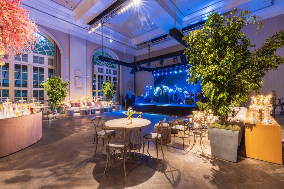 A spacious, elegantly decorated event hall with large arched windows allowing natural light to illuminate the interior. The polished wooden floor is clean and shiny, reflecting the ambient lighting. Several round tables with white tablecloths and small floral centerpieces are arranged throughout the room, complemented by stylish chairs. Large potted trees and floral arrangements add a touch of greenery to the space. In the background, a stage is set up with professional lighting and a live band performing, creating a lively atmosphere. The room's surfaces, including the wooden floors, table surfaces, and glass windows, are shown to be well-maintained, dust-free, and gleaming, highlighting the high hygiene and cleanliness standards upheld by Maida Vale Cleaners during event preparations and post-event cleaning services.