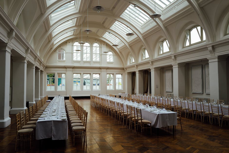 An elegant event space with polished wooden floors and high ceilings, featuring round tables decorated with tall glass vases filled with fresh pink and white flowers, accompanied by silver Chiavari chairs with cushioned seats. The room is well-lit with natural light from large windows, and the surfaces, including the tabletops and floors, appear spotless and well-maintained. A small side table holds a yellow floral arrangement, and soft beige drapes hang behind the tables. The overall scene reflects a deep cleaning and surface sanitisation tailored for event venues, with a focus on hygiene and visual appeal, typical for professional event cleaning services provided by Maida Vale Cleaners.