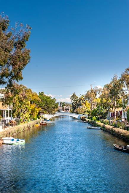 A scenic view of the canal in Little Venice, Maida Vale, with calm blue water reflecting the clear sky above. The canal is lined with residential houses, some with outdoor seating areas and lush greenery. Several small boats are anchored along the edges, and a small white pedestrian bridge spans the water in the background. Tall trees with green foliage frame the scene, adding to the tranquil atmosphere. The bright sunlight highlights the cleanliness and well-maintained appearance of the surroundings, aligning with the cleaning and maintenance theme of Maida Vale Cleaners' domestic cleaning services in the area.