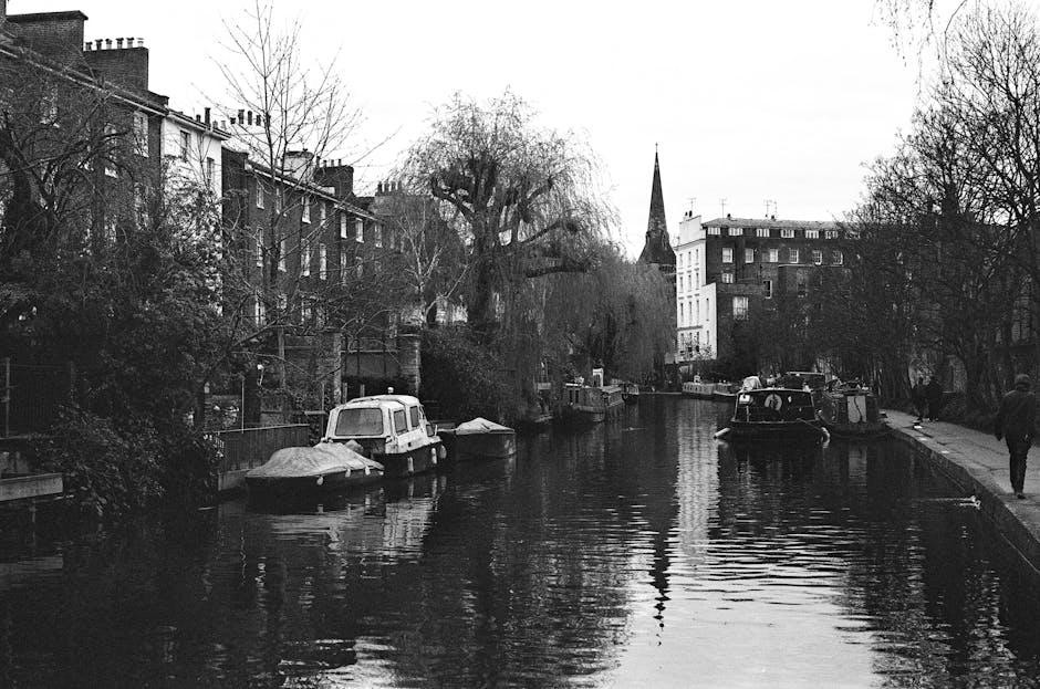 A black and white photograph showcases a serene scene along a residential canal in Maida Vale, with boats moored on both sides of the water. The canal features calm, reflective surface with visible ripples, bordered by paved walkways on each side. On the left, there are houses with brick facades, some partially obscured by leafless trees. The right side has a few pedestrians strolling, with trees lining the walkway. In the background, a church spire rises above the surrounding buildings, and the sky appears overcast, contributing to the monochrome, subdued atmosphere. This image reflects the tranquil, historic character of the Maida Vale canal area, emphasizing cleanliness and maintenance ideals associated with domestic and commercial surface cleaning, as promoted by Maida Vale Cleaners.