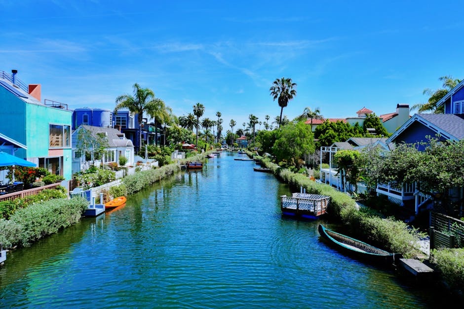 A scenic view of the canal in Little Venice, Maida Vale, with calm blue water reflecting the clear sky above. The canal is lined with residential houses, some with outdoor seating areas and lush greenery. Several small boats are anchored along the edges, and a small white pedestrian bridge spans the water in the background. Tall trees with green foliage frame the scene, adding to the tranquil atmosphere. The bright sunlight highlights the cleanliness and well-maintained appearance of the surroundings, aligning with the cleaning and maintenance theme of Maida Vale Cleaners' domestic cleaning services in the area.