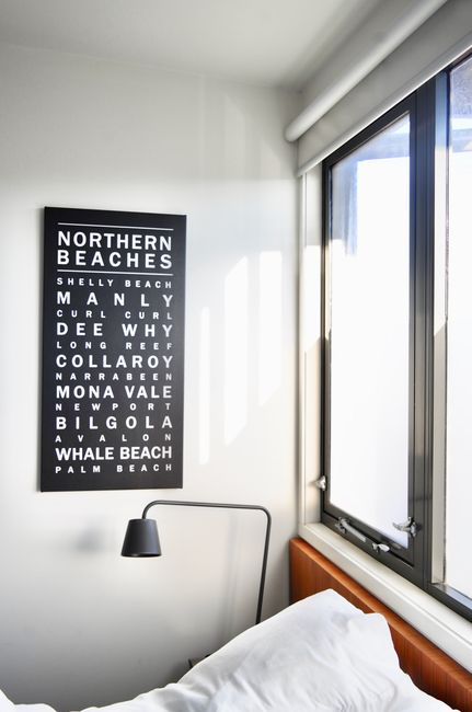 A portion of a bedroom featuring a white wall with a framed black and white typographic poster listing various beach name locations in the Northern Beaches area, including Shelly Beach, Manly, Curl Curl, Dee Why, Long Reef, Collaroy, Mona Vale, Newport, Balgowlah, Whale Beach, and Palm Beach. To the left of the poster, a sleek black adjustable reading lamp is mounted on the wall, positioned over a bed with white bedding and a wooden headboard. A large window with a white roller blind is on the right side, allowing natural light to illuminate the room, highlighting the smooth surfaces of the walls, window frame, and bedding, creating a clean and tidy appearance suitable for showcasing domestic cleaning and surface maintenance by Maida Vale Cleaners.