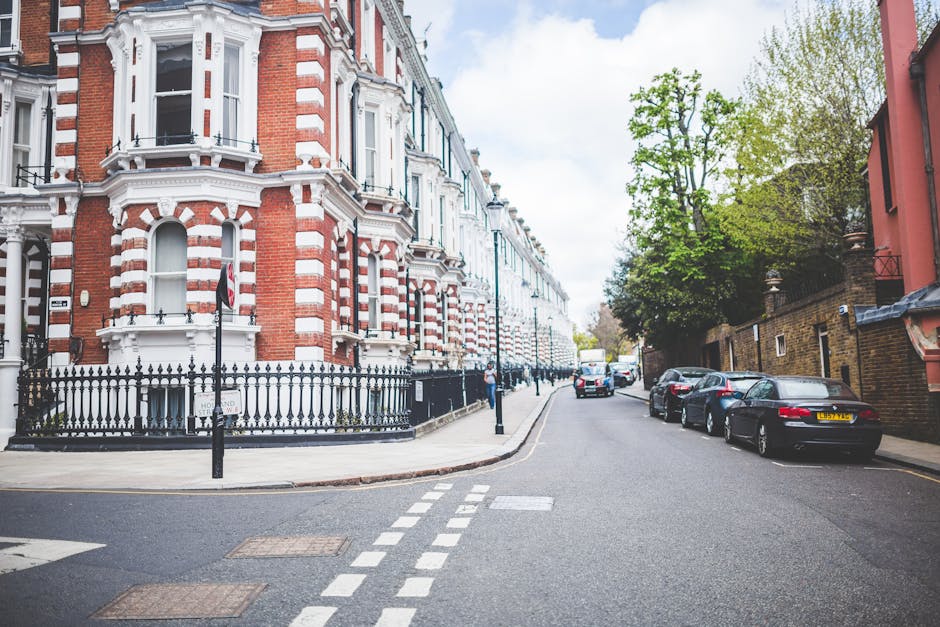 A street view of Maida Vale featuring a prominent Victorian-style building with red brick and white decorative accents, positioned at the corner of the street with a black wrought iron fence surrounding its base. The pavement is clean and well-maintained, with a striped pedestrian crossing visible in the foreground. Several parked cars are aligned along the right side of the street, including a black vehicle with a yellow UK license plate. Mature trees with green foliage line the sidewalk, adding greenery to the scene. Natural daylight illuminates the area, highlighting the tidy appearance of the street and the well-kept surfaces, reflecting the standards of cleanliness associated with professional domestic and commercial cleaning services by Maida Vale Cleaners, who specialise in surface cleaning, deep cleaning, and sanitisation for properties in this residential neighborhood.