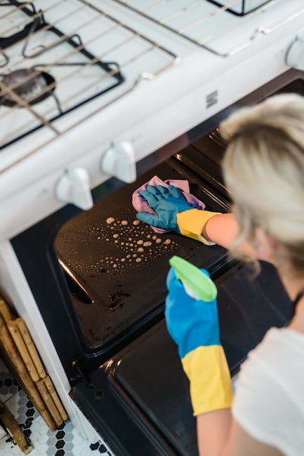 A person wearing yellow rubber gloves is cleaning the black stovetop of a white oven with a pink cloth, focusing on removing grease and spills. The oven door is open, revealing the dark interior, and the stovetop features metal grates and burner components. The surrounding area includes a white countertop and a tiled floor with a black and white pattern. The scene emphasizes surface cleaning and sanitisation, demonstrating thorough domestic cleaning practices, as provided by Maida Vale Cleaners, to maintain hygiene and shine in the kitchen environment.