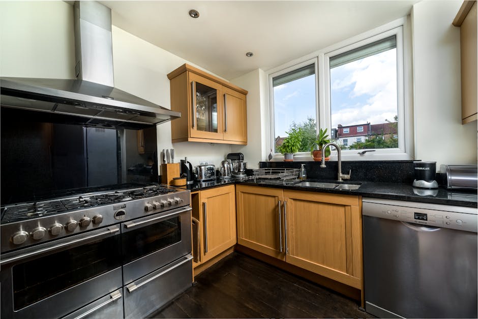 A modern kitchen with light wood cabinets and a black granite countertop, featuring a large stainless steel oven and stove with multiple burners on the left side. The countertop holds various small appliances including a toaster, kettle, and food processor, all neatly arranged. There is a black dish drying rack beside a stainless steel sink with a chrome faucet situated below a large window that lets in natural light, revealing a view of a suburban neighborhood with houses and greenery. The kitchen surfaces, including the stovetop and countertop, appear clean and well-maintained, indicating recent cleaning or regular sanitisation. Light-colored wooden cabinets above and below the counter provide ample storage, while the dark wooden floor adds contrast. The overall room is uncluttered, well-lit, and showcases a tidy, hygienic space suitable for domestic cleaning or deep cleaning services by Maida Vale Cleaners.