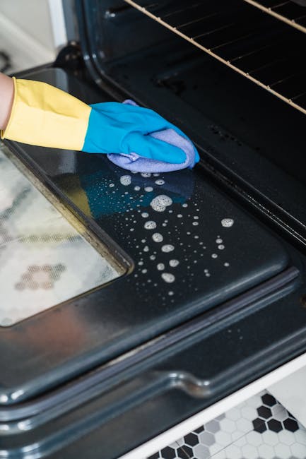 A person wearing yellow rubber gloves is cleaning the black stovetop of a white oven with a pink cloth, focusing on removing grease and spills. The oven door is open, revealing the dark interior, and the stovetop features metal grates and burner components. The surrounding area includes a white countertop and a tiled floor with a black and white pattern. The scene emphasizes surface cleaning and sanitisation, demonstrating thorough domestic cleaning practices, as provided by Maida Vale Cleaners, to maintain hygiene and shine in the kitchen environment.