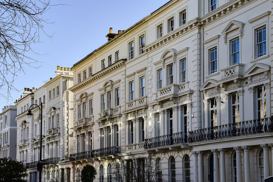 A row of elegant, white Victorian-style residential buildings with ornate architectural details, including decorative window moldings and black wrought iron balconies, situated along a street in Maida Vale. The facades are clean and well-maintained, with sunlight illuminating the intricate cornices and smooth surfaces. Lampposts line the sidewalk in front of the buildings, and leafless trees are visible in the foreground. The scene captures a tidy and picturesque residential area, highlighting the importance of surface cleaning and regular maintenance, as offered by Maida Vale Cleaners, in ensuring the aesthetic appeal and hygiene of properties in Sutherland Avenue.