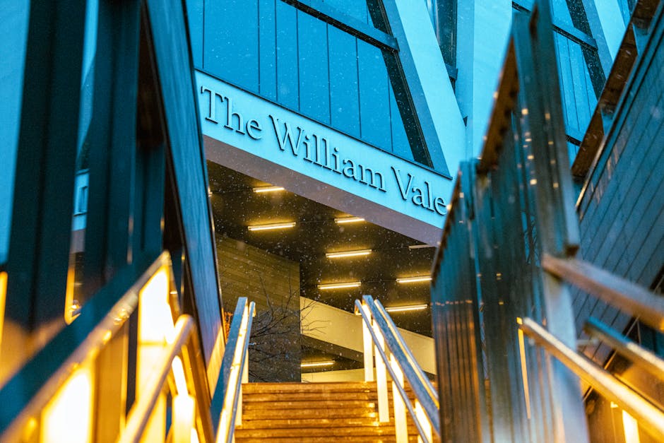 Exterior view of The William Vale building in Maida Vale, featuring modern dark-colored cladding with horizontal paneling. Illuminated signage displaying the building's name is visible above a staircase entrance. The staircase has wooden steps with metal handrails, and the area is lit by warm artificial lighting. Rain droplets can be seen on the building's surface, adding a reflective, wet appearance to the facade. The overall scene captures a sleek, contemporary architectural entrance during nighttime, emphasizing cleanliness and maintenance as part of the building's exterior presentation. Maida Vale Cleaners' branding is relevant to maintaining the building's pristine condition through professional cleaning services.