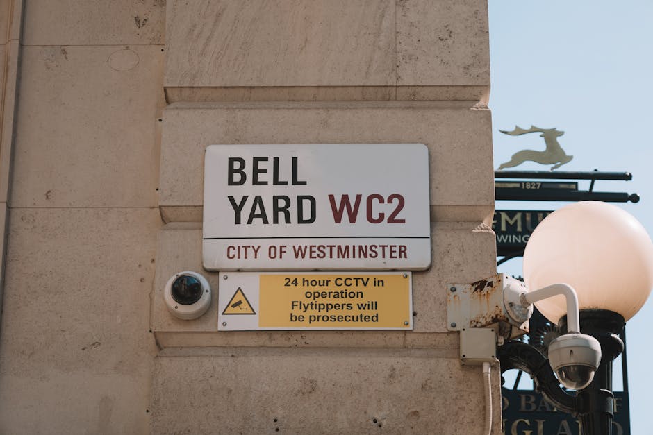 A close-up view of a beige stone wall featuring a rectangular street sign with black and red text that reads 'BELL YARD WC2 CITY OF WESTMINSTER'. Below the sign is a yellow notice indicating 24-hour CCTV operation and warnings about prosecution for flytipping. To the right, a classic black street lamp with a white, spherical light fixture is mounted on the wall. A security camera is also visible, affixed to the wall beneath the sign. The lighting appears natural, suggesting daylight, with the background showing a clear sky and a decorative sign with a silhouette of a stag. The scene emphasizes outdoor urban cleaning and security measures consistent with Westminster Council regulations, typical of Maida Vale.